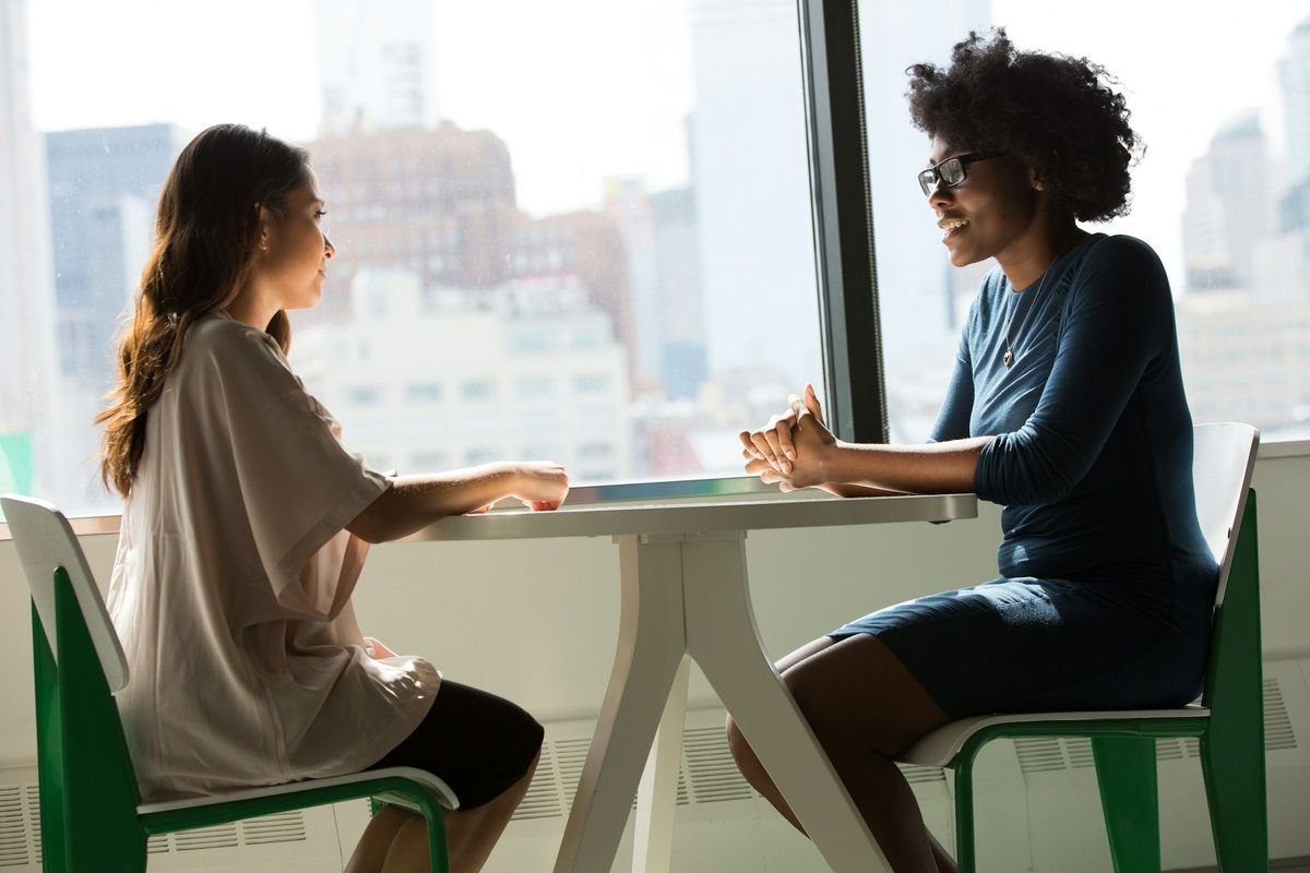Two women talk at a table by a sunny window.